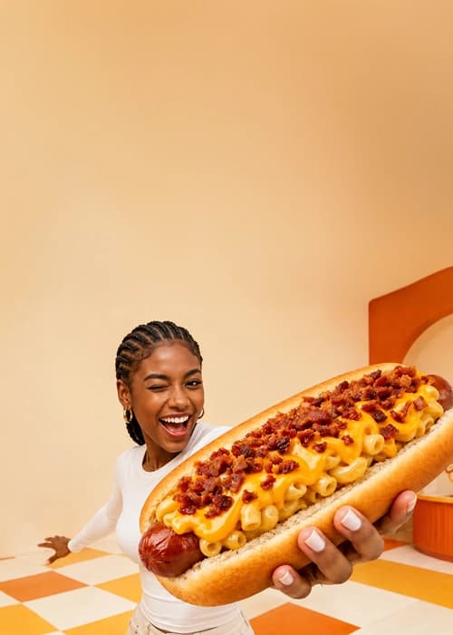 Smiling person holding out a giant mac and cheese dog topped with bacon crumbles in a retro diner