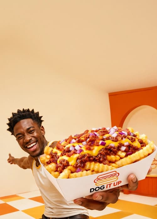 Smiling person holding out a giant loaded crinkle-cut fries with chili, cheese and onion in a retro diner