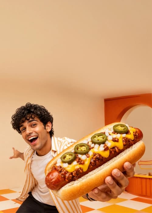 Smiling person holding out a giant chili cheese dog topped with jalapenos and onion in a retro diner